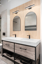 A Modern Bathroom With White Oak Wood Slat Paneling On The Wall With Arched Mirrors, Mounted Lights, And A Wooden Cabinet.