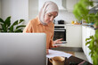 © Johnér - Smiling woman with hijab using cell phone during work from home in kitchen