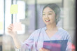 © David - Young Creative business asian woman writing on sticky notes on a glass wall, female colleague looking.