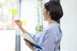 © David - Young Creative business asian woman writing on sticky notes on a glass wall, female colleague looking.