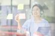 © David - Young Creative business asian woman writing on sticky notes on a glass wall, female colleague looking.