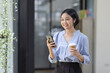 © David - Young beautiful Asian woman holding coffee paper cup and looking at smartphone while sitting at workplace. Happy university student girl using mobile phone. Business woman drinking coffee and smiling.
