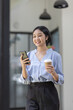© David - Young beautiful Asian woman holding coffee paper cup and looking at smartphone while sitting at workplace. Happy university student girl using mobile phone. Business woman drinking coffee and smiling.