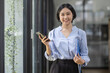 © David - Young beautiful Asian woman holding coffee paper cup and looking at smartphone while sitting at workplace. Happy university student girl using mobile phone. Business woman drinking coffee and smiling.