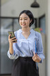 © David - Young beautiful Asian woman holding coffee paper cup and looking at smartphone while sitting at workplace. Happy university student girl using mobile phone. Business woman drinking coffee and smiling.