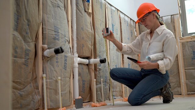 Woman architect inspecting toilet plumbing in a new home construction using a touchpad tablet and measuring tape.