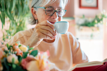 Sticker - Blurred senior woman sitting at cafe table reading a book or studying enjoying an espresso coffee. Elderly lady holding a white coffee cup