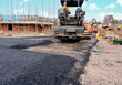 Asphalt paver filled with hot tarmac laying new road surface on new residential housing development site and roadworker operator in orange hi-viz next to it