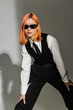 © LIGHTFIELD STUDIOS - youthful and expressive asian woman with dyed red hair posing in dark sunglasses, black and white business casual clothes and looking at camera on grey shaded background, generation z