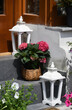 A composition of hydrangea in a wicker basket, standing between two white wooden lanterns on granite steps in front of the entrance to the flower shop, on the background of a wall, a window and a door
