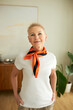 Confident elderly blond woman fashion stylist standing in living-room in white t-shirt and orange and black silk scarf around her neck, looking at camera with smile putting hands in pockets