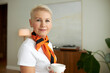 © Anatoliy Karlyuk - Attractive elderly female teacher or dean of university posing against cozy classroom with white mug in hands, going to have some coffee during break, looking at camera with positive facial expression
