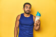 Athletic young Latino man holding a protein shake against a yellow background shrugs shoulders and open eyes confused.