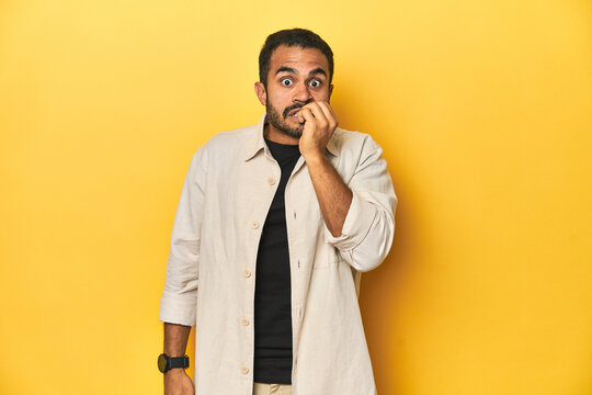 Casual young Latino man against a vibrant yellow studio background, biting fingernails, nervous and very anxious.