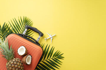 Unwind in summer paradise. From top view, orange suitcase, miniature plane, coast food like fresh coconut, pineapple and palm leaves arranged on yellow background, inviting space for text or advert