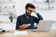 © sofiko14 - Feverish bearded man in casual clothes touching forehead while looking at laptop camera in dining room of apartment. Unwell arabian manager arranging virtual appointment with health professional.