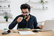 © sofiko14 - Young indian businessman tasting fresh greens in vegan meal while sitting at office desk in bright apartment. Self-employed worker enjoying nutritious food during lunch hour while telecommuting.