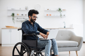 Wall Mural - Joyful indian man in wheelchair holding laptop on knees while staying in middle of bright open-plan kitchen. Happy young adult surfing internet webpages while recovering after accident at home.