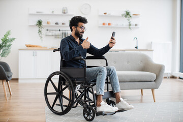 Wall Mural - Cheerful wheelchair user in spectacles and casual outfit utilizing phone webcam while enjoying free time indoors. Bearded indian male receiving online conference call via mobile application about job.