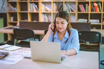 Wall Mural - Business asian woman sitting at her desk with laptop using mobile phone chatting with customers or giving financial advice.