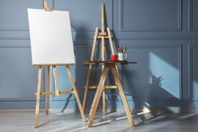 Easel With Blank Canvas And Different Art Supplies On Wooden Table Near Grey Wall Indoors