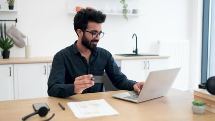 Wall Mural - Attractive bearded man in spectacles holding blank credit card while typing on portable computer in kitchen interior. Happy arabian gentleman doing online banking transaction while working remotely.