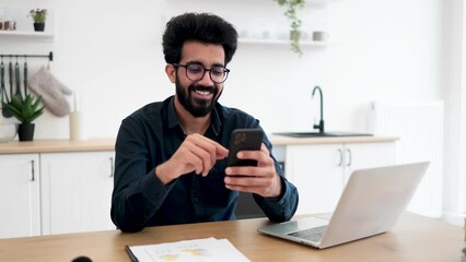 Wall Mural - Positive indian male in spectacles holding smartphone while staying in modern workplace at home. Efficient freelance worker texting message to colleague while performing project for employer indoors.