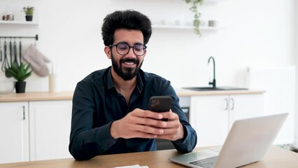 Wall Mural - Positive indian male in spectacles holding smartphone while staying in modern workplace at home. Efficient freelance worker texting message to colleague while performing project for employer indoors.