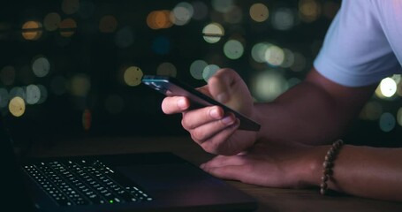 Close-up of business man writing a message on a mobile phone while working on a laptop on background of a night city. Male businessman uses a mobile phone while working at a computer in the evening