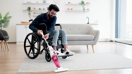 Wall Mural - Helpful young man with disability holding cordless vacuum cleaner while sitting in wheelchair in open-plan kitchen. Active person in casual outfit collecting dust and dirt from carpet in apartment.