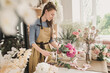 woman florist in beige apron is working in  flower shop. Floral design studio, making decorations and arrangements. Flower delivery, order creation. Small business.