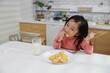 © feeling lucky - Kid asian black hair girl sitting on table and drinking milk while eating vanilla cookies for breakfast with enjoying time. Tasty food and delicious food with happy meal lifestyle kid concept