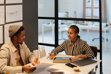 Aufkleber - African American guy with papers looking at young female colleague using tablet while both sitting by desk with gadgets and office supplies