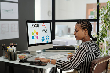 Aufkleber - Side view of young serious African American female webdesigner sitting by workplace in front of computer monitor and using tablet