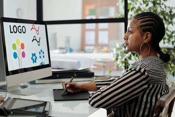 Aufkleber - Serious African American female webdesigner sitting by desk in front of computer monitor with logo on screen and working over new project