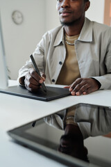 Aufkleber - Close-up of young black man sitting by workplace in front of computer screen in office and drawing graphic sketches or retouching images