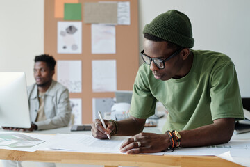 Poster - Young African American designer or architect in casualwear and eyeglasses drawing sketch of new building in office while bending over papers