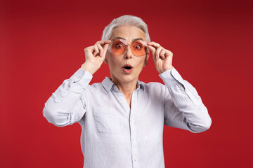 Wall Mural - Excited gray haired woman wearing sunglasses, looking at camera isolated on red background