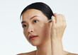 © Wesley JvR/peopleimages.com - Asian woman, dropper and keratin on face for beauty, skincare or cosmetics against white studio background. Happy female model applying oil drop to skin for hydration, moisturizer or facial treatment