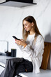 Happy young woman relaxing at home she is sitting on counter kitchen and using mobile smartphone