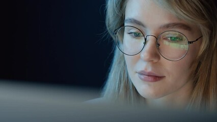 Panning right close up of cheerful woman sitting in front of computer at night and choosing something to watch wearing glasses with screen reflection in them