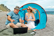 © Caterina Trimarchi - Couple having fun, drinking wine and talking with friends in video call in laptop in picnic near tent on seaside. Vacation summer on the beach in outdoors. Travel, vacation and friendship concept.