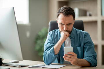 Stressed businessman looking at smartphone screen while sitting at desk in office