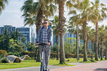 Young man riding electric scooter eco friendly transport along the promenade in the city with palm trees. Male driving e-scooter sustainable and outdoor in summer day. Sustainable lifestyle