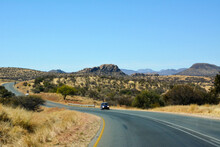 A Car Drives Along A Long Winding Road In Perspective In A Desert Area. Small Mountains Are In The Distance