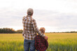 © Acronym - Farmer and his son in front of a sunset agricultural landscape. Man and a boy in a countryside field. Fatherhood, country life, farming and country lifestyle concept.