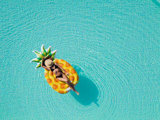 Wall Mural - Sexy young female in swimsuit bikini, in a straw hat and sunglasses floating on blue swimming pool waves on giant inflatable Yellow Pineapple tube. Chill out summer vacation in luxury resorts concept