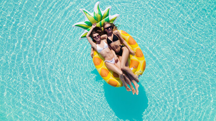 Wall Mural - Top view of two girls relaxing on inflatable big pineapple in the pool. Aerial view of ladies relaxing on the floating mattress.