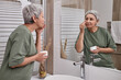 © Seventyfour - Side view portrait of senior Asian woman looking in mirror and using facial cream in anti aging routine
