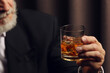 © New Africa - Man holding glass of whiskey with ice cubes on dark background, closeup